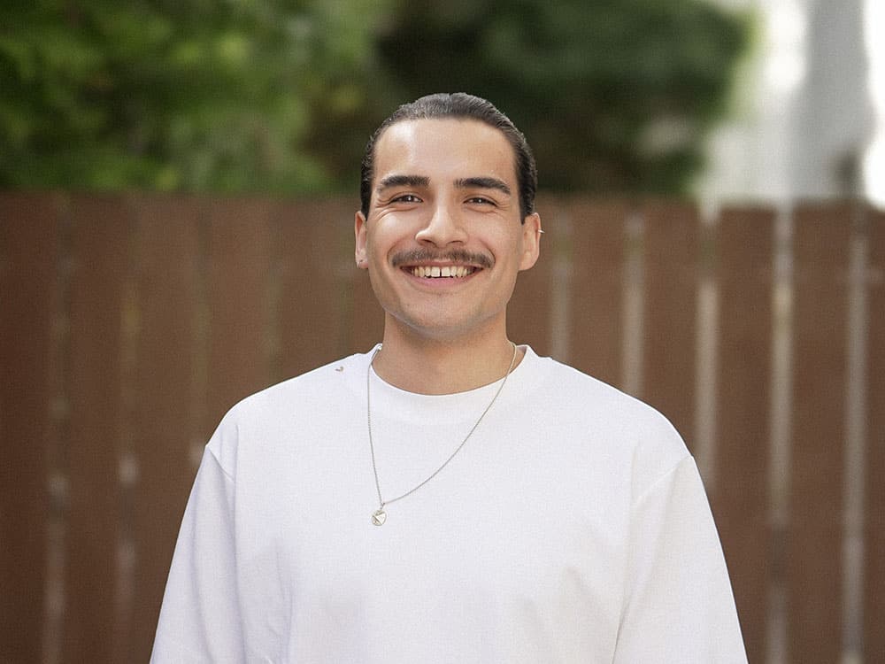 A young man with a mustache smiles in a blurred green background with a wooden fence.